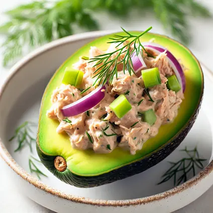 Halved avocados stuffed with tuna salad, garnished with red onion, celery, and fresh dill on a white plate.